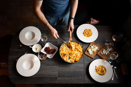 Man sets the table, homemade pasta, bacon, feta and tomatoes, cheeses with blue mold, wine. Deep plates for pasta. Top view.の写真素材