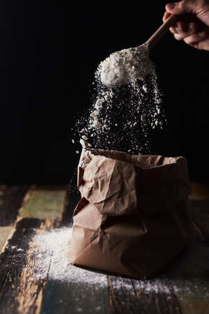 Wheat flour in a craft package. Freeze movement, spoon dark background, sprinkle flour. Front view.の写真素材