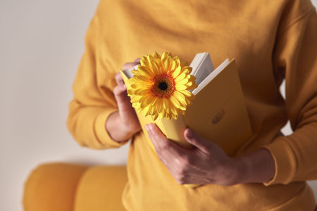 A yellow book with a flower bookmarked in the hands of a woman on a yellow sweater background. Front...