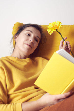 A beautiful girl dreams of sitting in a yellow chair with a book and a flower in her hands. Front...