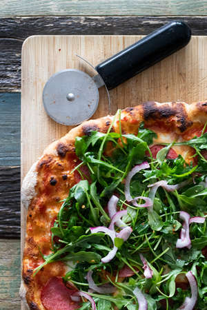 Homemade pizza with tomato sauce, sausage and arugula salad with onions on a wooden board with a knife. Top view.の写真素材