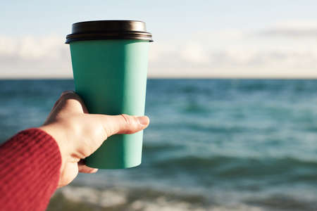A woman holds a disposable paper glass with coffee by the sea. Front view.の写真素材