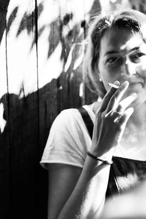 Black and white portrait of a beautiful woman against the background of a wooden wall. A woman smokes a cigarette. Vertical photo.の写真素材