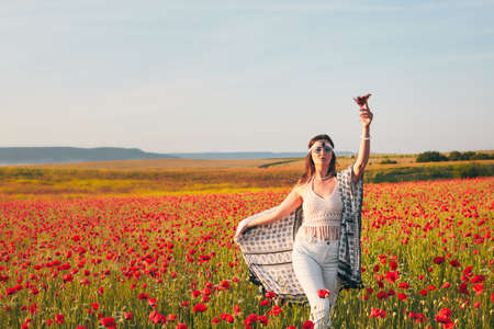 A young woman poses in a field with red wild poppies at sunset. She is holding a glass of strawberries. Front view.の写真素材