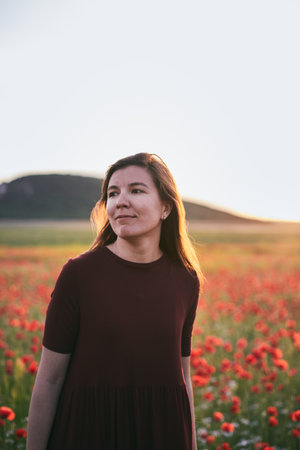 A young woman poses in a field with red wild poppies at sunset. Vertical photo.の写真素材