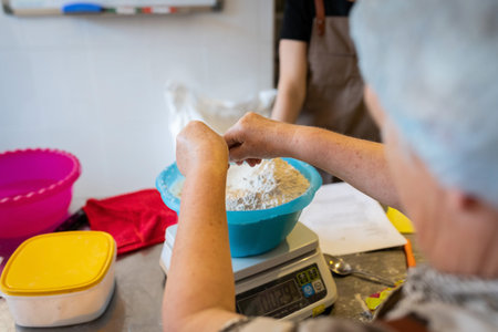 A mature woman measures ingredients for baked goods in the kitchen of a small bakery. Front view.