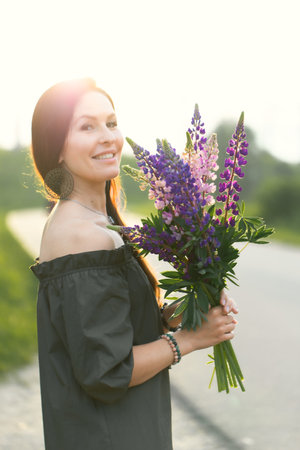 Attractive woman on the road in a summer field among Lupinus flowers. Vertical photo.の写真素材