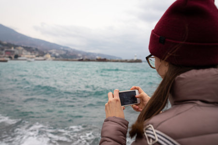 A woman takes pictures on her phone of waves in the sea on the shore of Yalta. Front view.の写真素材