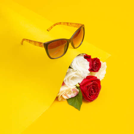 A bouquet of blooming red and white roses protrudes from under the yellow paper. There are glasses above the flowers. An abstract creative idea. Valentine's Day. Summer composition, sunny backgrounds.の写真素材