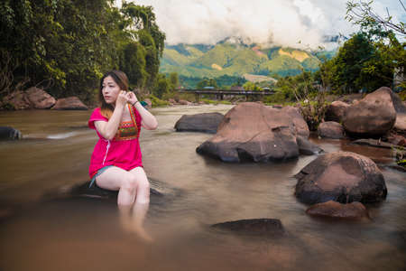 A beautiful woman wearing a pink dress sitting on the rocks in a river is adjusting her earrings amid nature, with a mountainous background and clouds.の写真素材
