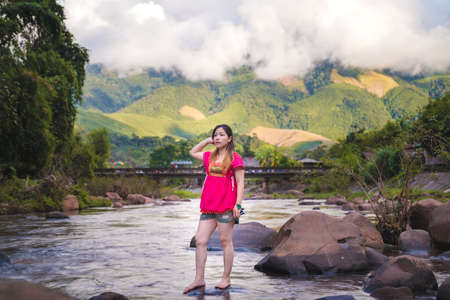 Beautiful women wearing pink outfits standing on rocks in a stream surrounded by nature, with a background of mountains and clouds.の写真素材