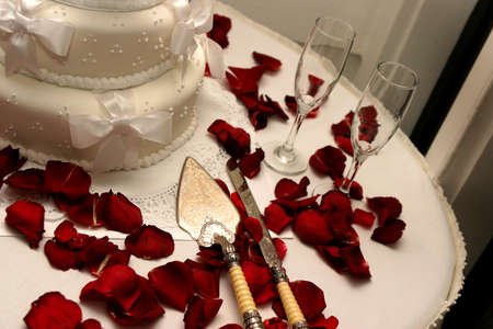 white wedding cake with red rose peddles covering the table along with decrative cake knifes,の写真素材