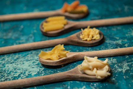 Various mix of dry pasta on wooden spoons. Dry pasta on rustic blue background. Diet and food concept.の写真素材