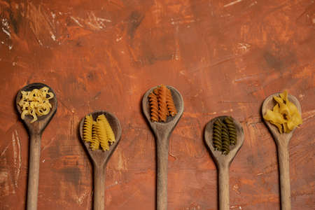 Various mix of dry pasta on wooden spoons. Dry pasta on rustic brown background. Top view with copy space. Diet and food concept.の写真素材