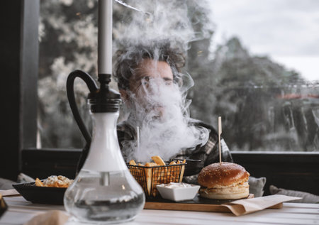 Handsome boy eating at a restaurant while smoking shisha. Surrounded with smoke.の写真素材