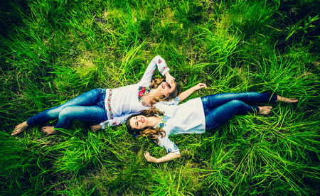 Two pretty girls lying on the fresh green grass in embroidery shirtsの写真素材