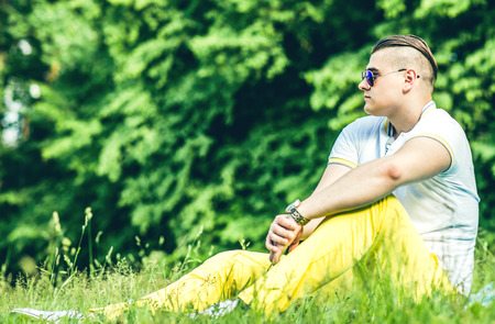Pretty young man in sunglasses sittin on the grass in the parkの写真素材
