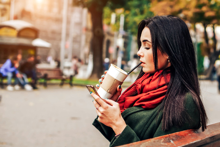 Brunette girl sitting in the park with coffee and phoneの写真素材