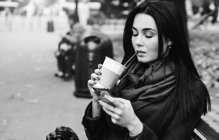 Brunette girl sitting in the park with coffee and phoneの写真素材