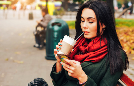Brunette girl sitting in the park with coffee and phoneの写真素材
