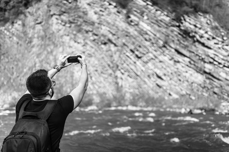 Traveler with backpack making photo on mobile phone near mountain river, black and whiteの写真素材