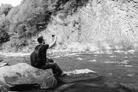 Traveler with backpack making photo on mobile phone near mountain river, black and whiteの写真素材
