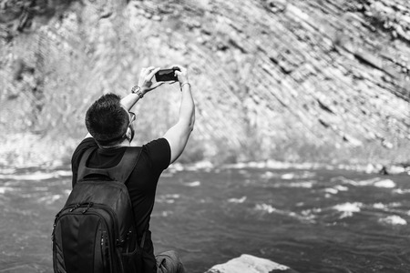 Traveler with backpack making photo on mobile phone near mountain river, black and whiteの写真素材