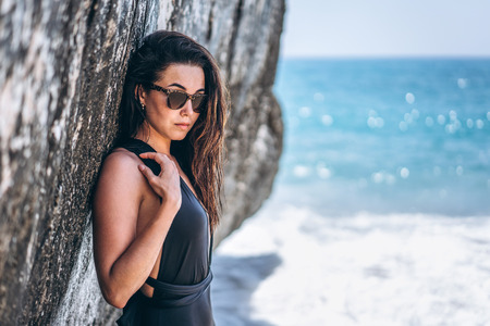 Pretty brunette pan asian girl in black swimsuit relaxing under the rock on the beachの写真素材