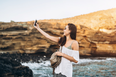 Pretty long hair tourist girl relaxing on the stones near sea with phoneの写真素材