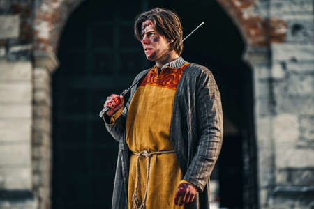 Portrait of a young medieval warrior in armor with sword on his shoulder and wounds on the face. In the process of battle, near the castle.の写真素材