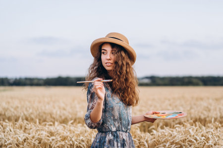 Closeup portrait of a beautiful young woman with curly hair. Woman in dress and hat standing in wheat field.の写真素材