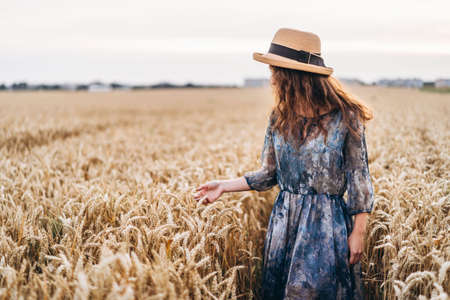 Closeup portrait of a beautiful young woman with curly hair. Woman in dress and hat standing in wheat field.の写真素材
