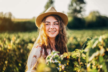 Portrait of a beautiful young woman with curly hair and freckles face. Woman in dress and hat posing in wheat field at sunset and looking at camera.の写真素材