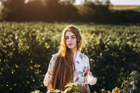 Portrait of a beautiful young woman with curly hair and freckles face. Woman in dress and hat posing in wheat field at sunset and looking at camera.の写真素材