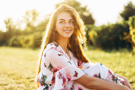 Portrait of a beautiful young woman with curly hair and freckles face. Woman in dress and hat posing in wheat field at sunset and looking at camera.の写真素材