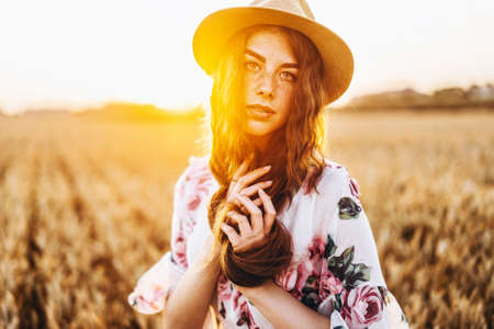Portrait of a beautiful young woman with curly hair and freckles face. Woman in dress and hat posing in wheat field at sunset and looking at camera.の写真素材