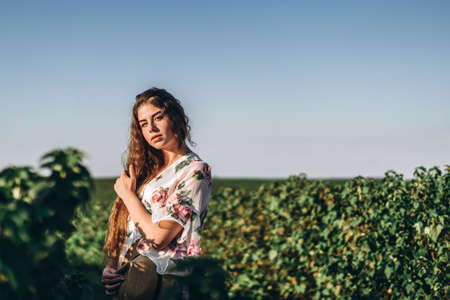 Portrait of a beautiful young woman with curly hair and freckles face. Woman in dress and hat posing in wheat field at sunset and looking at camera.の写真素材