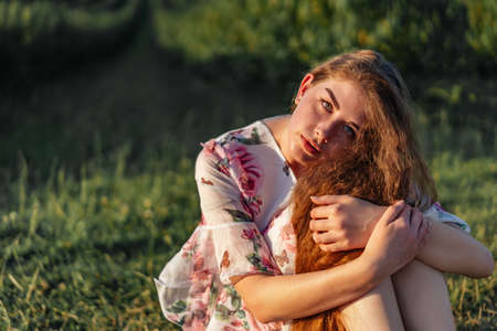 Portrait of a beautiful young woman with curly hair and freckles face. Woman in dress and hat posing in wheat field at sunset and looking at camera.の写真素材