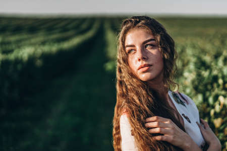 Portrait of a beautiful young woman with curly hair and freckles face. Woman in dress and hat posing in wheat field at sunset and looking at camera.の写真素材