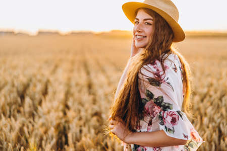 Portrait of a beautiful young woman with curly hair and freckles face. Woman in dress and hat posing in wheat field at sunset and looking at camera.の写真素材