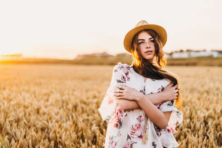 Portrait of a beautiful young woman with curly hair and freckles face. Woman in dress and hat posing in wheat field at sunset and looking at camera.の写真素材