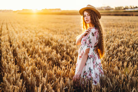 Portrait of a beautiful young woman with curly hair and freckles face. Woman in dress and hat posing in wheat field at sunset and looking at camera.の写真素材