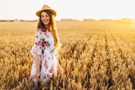 Portrait of a beautiful young woman with curly hair and freckles face. Woman in dress and hat posing in wheat field at sunset and looking at camera.の写真素材