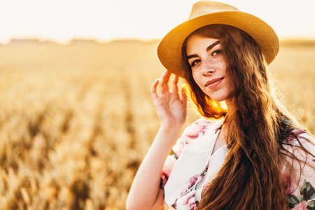 Portrait of a beautiful young woman with curly hair and freckles face. Woman in dress and hat posing in wheat field at sunset and looking at camera.の写真素材