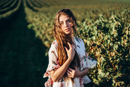 Portrait of a beautiful young woman with curly hair and freckles face. Woman in dress and hat posing in wheat field at sunset and looking at camera.の写真素材