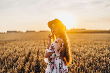 Portrait of a beautiful young woman with curly hair and freckles face. Woman in dress and hat posing in wheat field at sunset and looking at camera.の写真素材