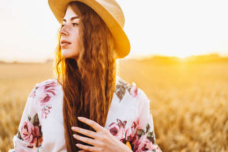 Portrait of a beautiful young woman with curly hair and freckles face. Woman in dress and hat posing in wheat field at sunset and looking at camera.の写真素材