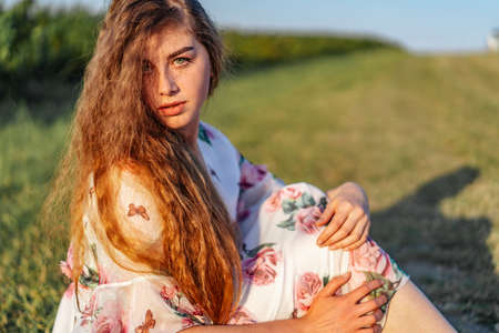 Portrait of a beautiful young woman with curly hair and freckles face. Woman in dress and hat posing in wheat field at sunset and looking at camera.の写真素材