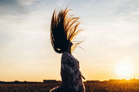 Portrait of a beautiful young woman with curly hair and freckles face. Woman in dress and hat posing in wheat field at sunset and looking at camera.の写真素材