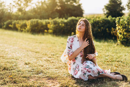 Portrait of a beautiful young woman with curly hair and freckles face. Woman in dress and hat posing in wheat field at sunset and looking at camera.の写真素材